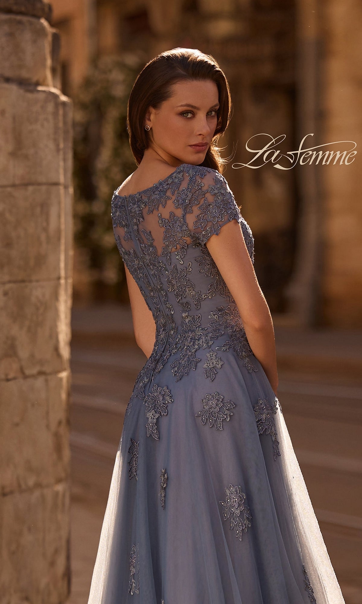 A woman in a blue Long Formal Dress: La Femme 29380 with lace appliques and floral embroidery stands outdoors, looking over her shoulder.